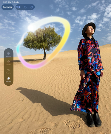iPad Pro, parte frontal, en posición vertical, negro espacial, esquinas redondeadas, la pantalla muestra la imagen de una mujer con un vestido de colores de pie en un desierto con un árbol de fondo, rodeado por un círculo de colores que muestra la capacidad de Apple Intelligence para borrar objetos del fondo