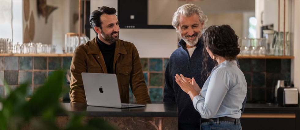 Dos hombres y una mujer están conversando, y se ve una MacBook sobre una mesa.
