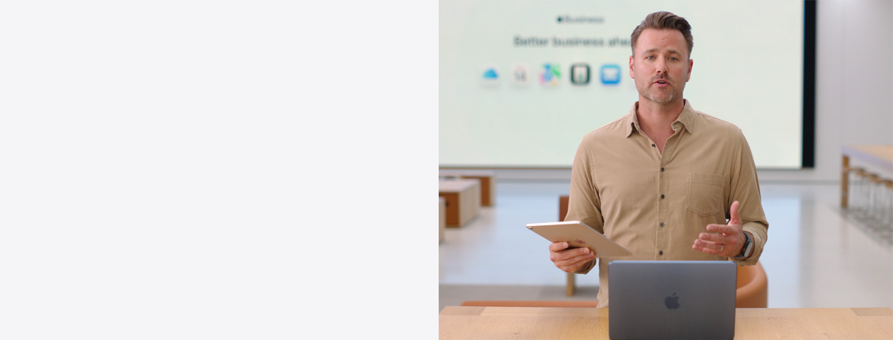 Moderator spricht und hält dabei ein iPad in der Hand, vor ihm steht ein MacBook auf einem Tisch, während ein großes Display hinter ihm verschiedene Business App Symbole anzeigt.