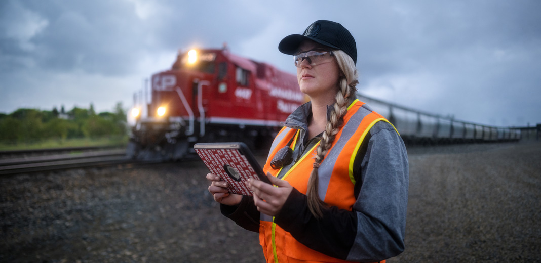 Eine Bahnmitarbeiterin hält ein iPad in der Hand und schaut auf die Gleise, während ein Zug vorbeifährt