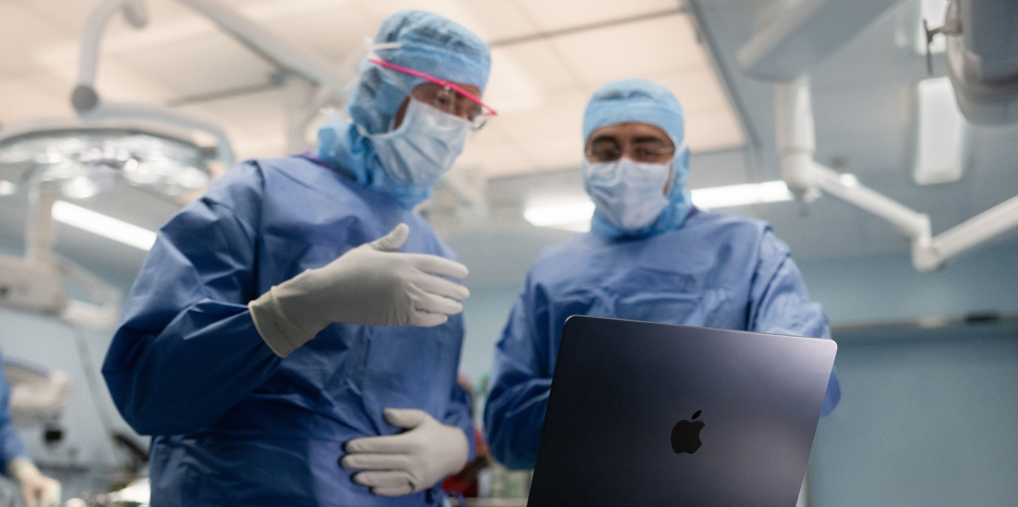 Zwei Chirurgen im Operationssaal schauen auf ein MacBook, wobei einer eine Geste mit der Hand macht, während der andere beobachtet
