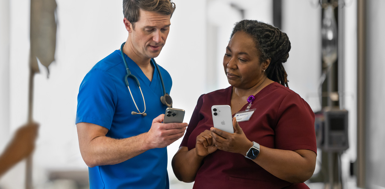 Nam nhân viên y tế mặc đồng phục màu xanh dương đang chỉ điều gì đó trên iPhone của mình cho một nữ nhân viên y tế mặc đồng phục màu đỏ, cũng đang cầm một chiếc iPhone.