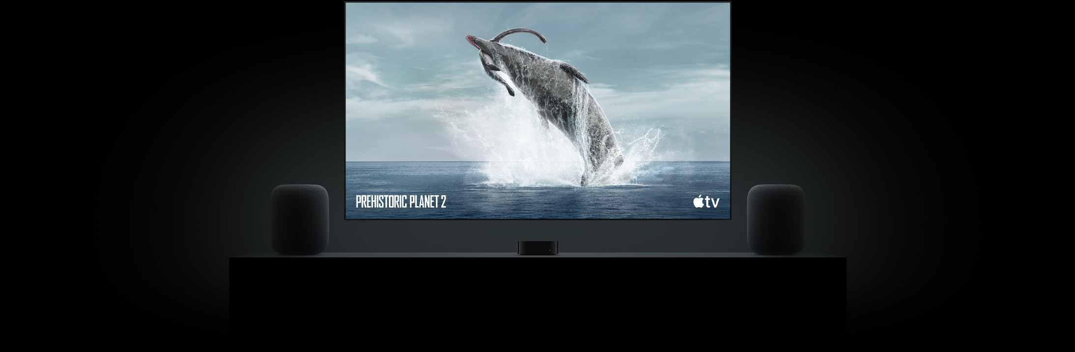 Large flat screen TV featuring a vivid image of a dinosaur from Prehistoric Planet. The TV is hung above an Apple TV 4K and framed by two HomePod speakers placed on a living room console