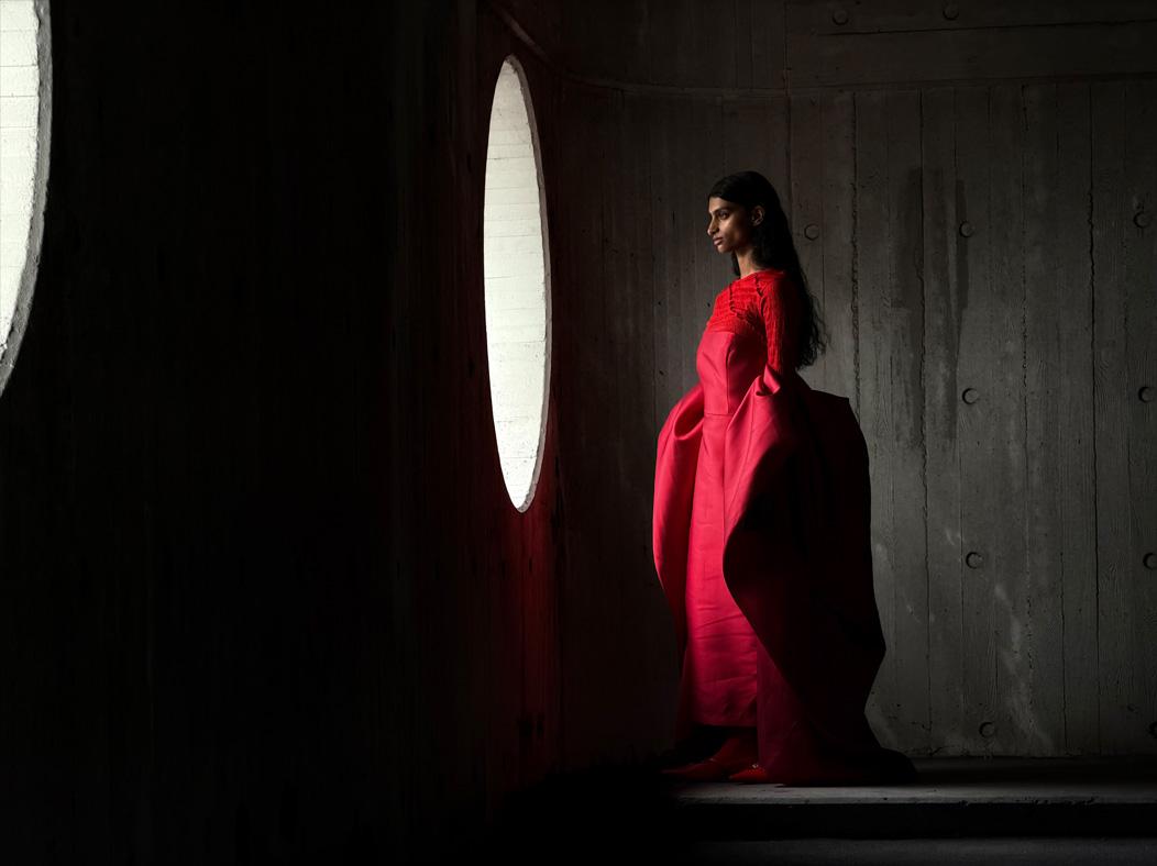 Una mujer con vestido largo rojo está siendo fotografiada de pie en una sala vacía. La distancia focal de la imagen avanza en la app Fotos para demostrar las capacidades del sistema de cámaras Pro, se muestra una distancia focal de 100 mm