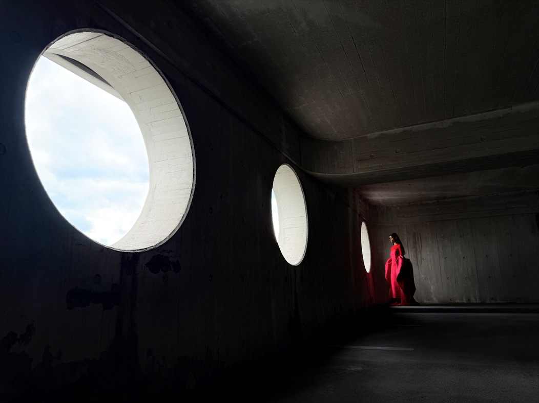 Una mujer con vestido largo rojo está siendo fotografiada de pie en una sala vacía. La distancia focal de la imagen avanza en la app Fotos para demostrar las capacidades del sistema de cámaras Pro, se muestra una distancia focal de 24 mm