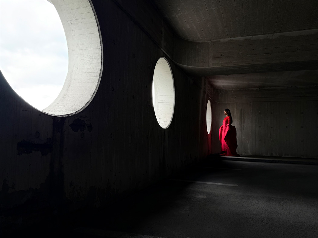 Una mujer con vestido largo rojo está siendo fotografiada de pie en una sala vacía. La distancia focal de la imagen avanza en la app Fotos para demostrar las capacidades del sistema de cámaras Pro, se muestra una distancia focal de 28 mm