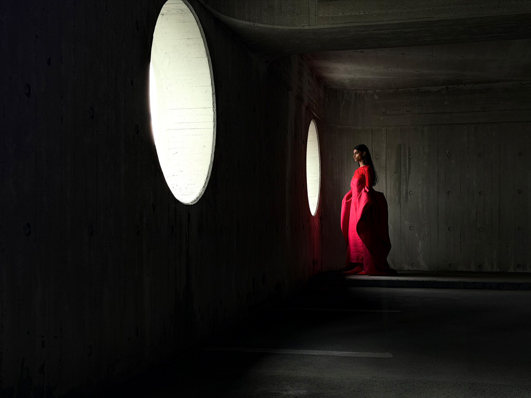 Una mujer con vestido largo rojo está siendo fotografiada de pie en una sala vacía. La distancia focal de la imagen avanza en la app Fotos para demostrar las capacidades del sistema de cámaras Pro, se muestra una distancia focal de 48 mm