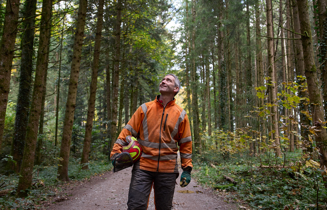 Ein Mann in einer orangefarbenen Warnjacke hält einen Schutzhelm und steht auf einem Waldweg in einem nachhaltig bewirtschafteten Wald in der Schweiz