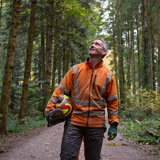 Ein Mann in einer orangefarbenen Warnjacke hält einen Schutzhelm und steht auf einem Waldweg in einem nachhaltig bewirtschafteten Wald in der Schweiz