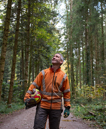 Ein Mann in einer orangefarbenen Warnjacke hält einen Schutzhelm und steht auf einem Waldweg in einem nachhaltig bewirtschafteten Wald in der Schweiz