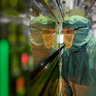 Eine Mitarbeiterin in vollständiger Schutzkleidung blickt durch ein Sichtfenster in eine Halbleitermaschine