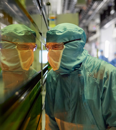 Eine Mitarbeiterin in vollständiger Schutzkleidung blickt durch ein Sichtfenster in eine Halbleitermaschine